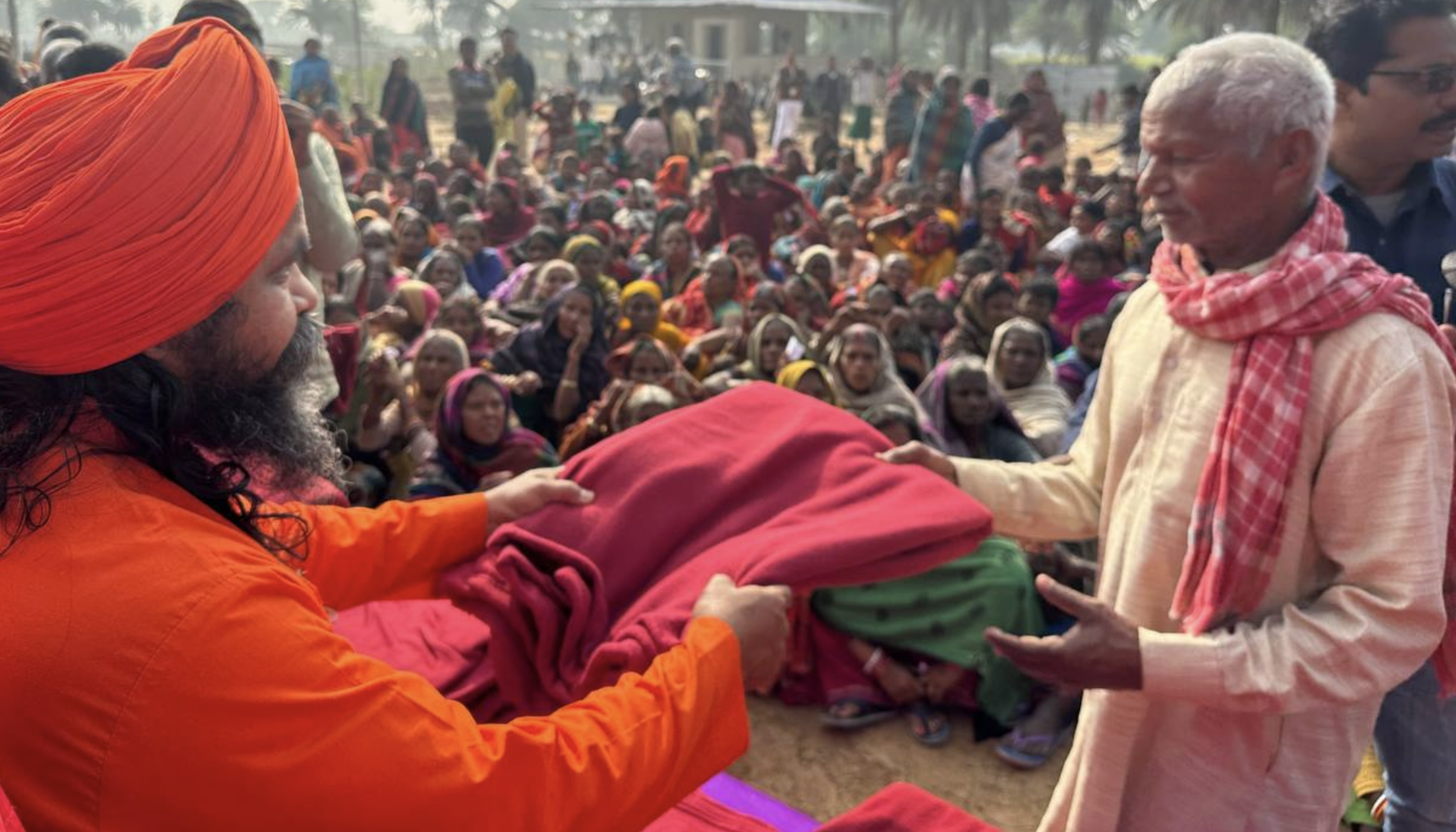 An Indian Yogi with an orage turban and robe handing a blanket to an an older Indian man with a crowd in the background.
